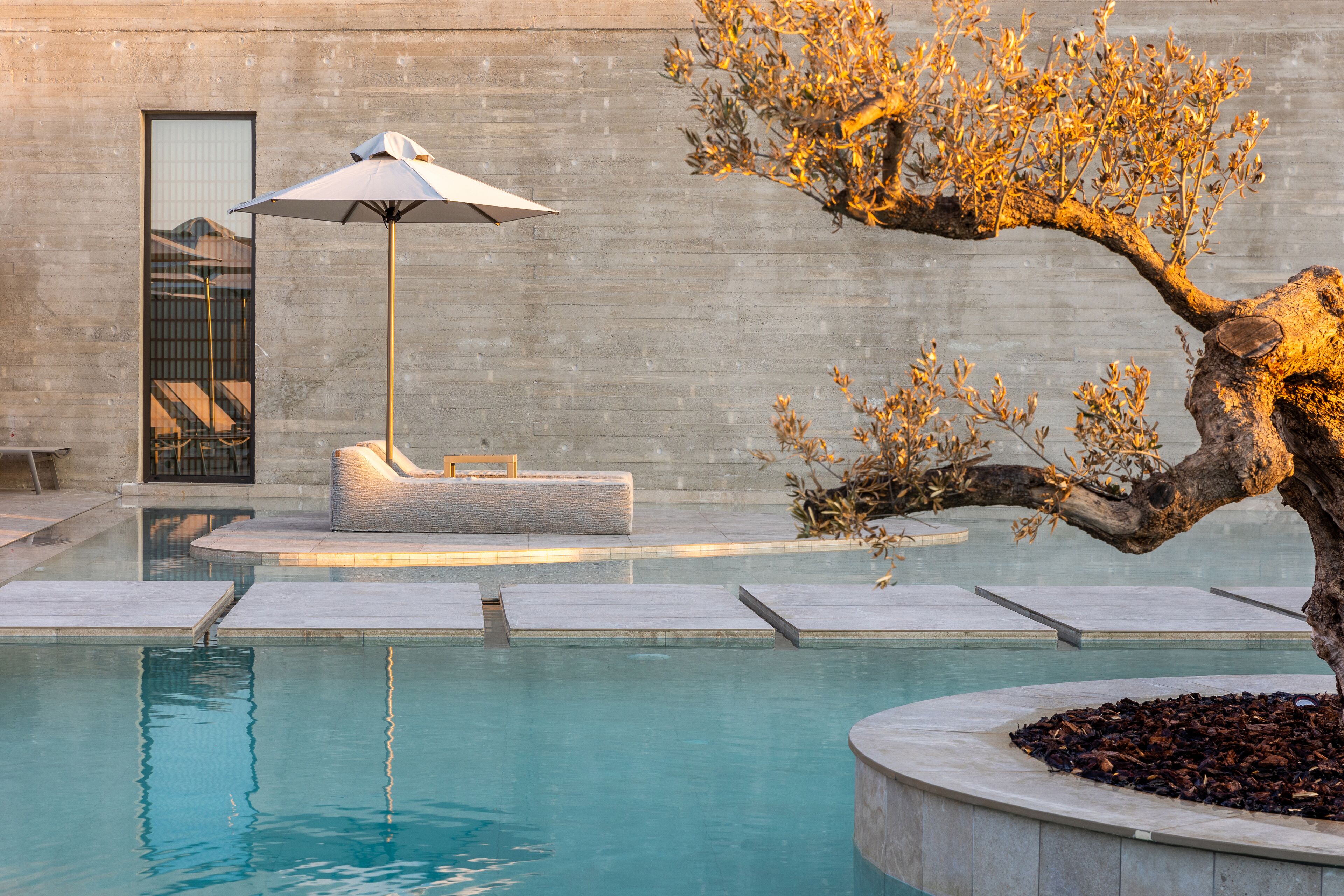 Un área de piscina con una silla de descanso y una sombrilla, un árbol y un muro de concreto.