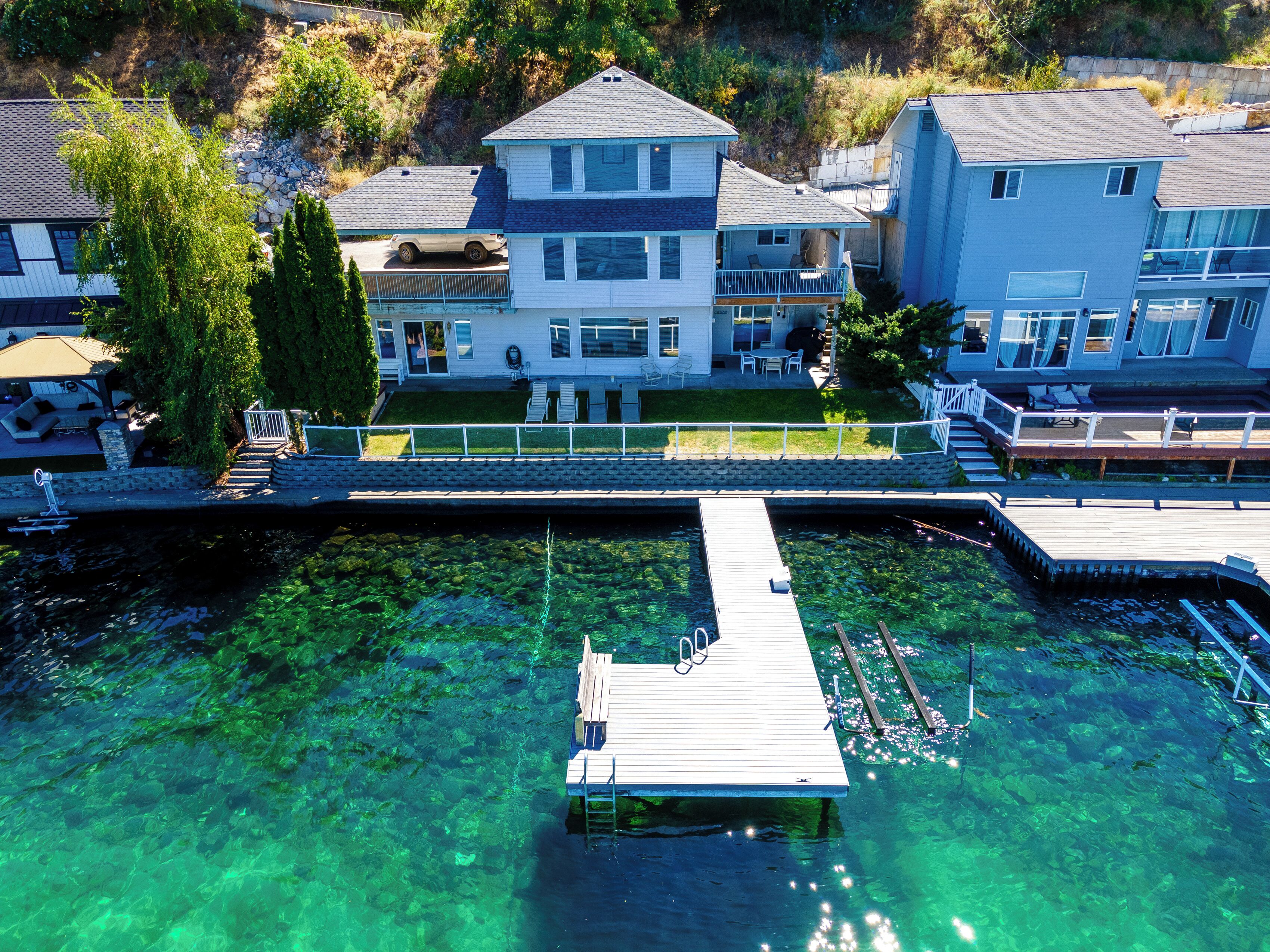 A house with a dock extending into a clear lake, surrounded by greenery and other houses.