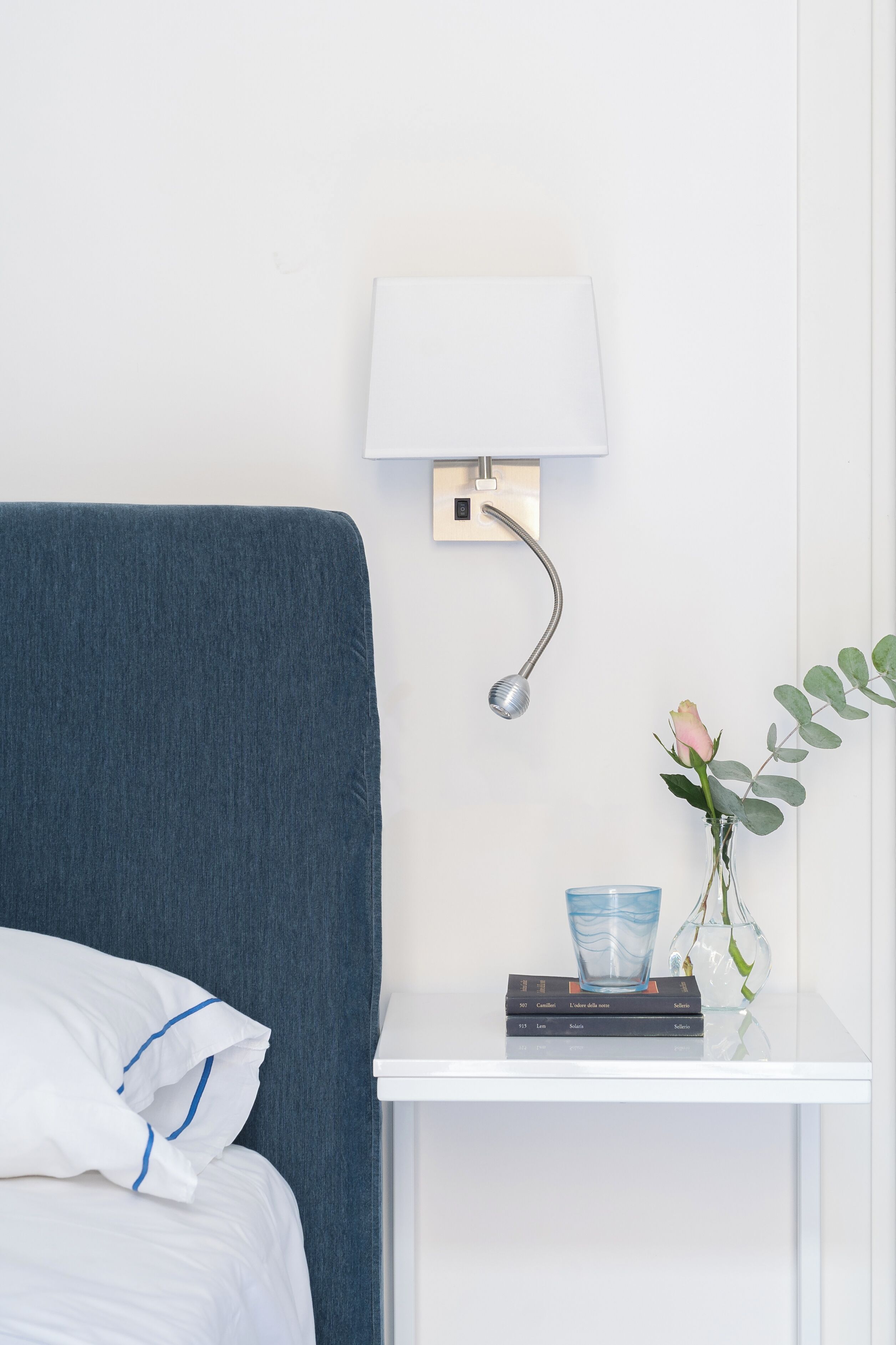 A neatly made bed with a blue headboard, a bedside table with a lamp, a vase with a rose, and a stack of books.