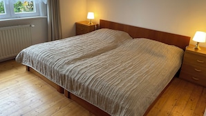 A bedroom with a wooden bed, two bedside tables with lamps, a window with curtains, and a radiator.