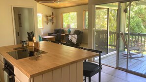 A modern kitchen with a central island, a dining area with a sofa and chairs, and a sliding glass door leading to a balcony with a view of greenery. - Seaside House, a Peaceful 2 Bedroom Retreat in walking distance to Kehena Beach (Pahoa)