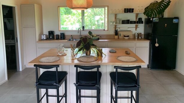 A modern kitchen with a central island, black bar stools, and a large window with greenery outside. - Seaside House, a Peaceful 2 Bedroom Retreat in walking distance to Kehena Beach (Pahoa)
