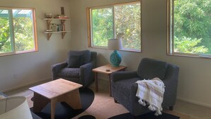 A cozy room with two armchairs, a small wooden table, and a lamp on a side table. Large windows provide views of greenery outside. - Seaside House, a Peaceful 2 Bedroom Retreat in walking distance to Kehena Beach (Pahoa)