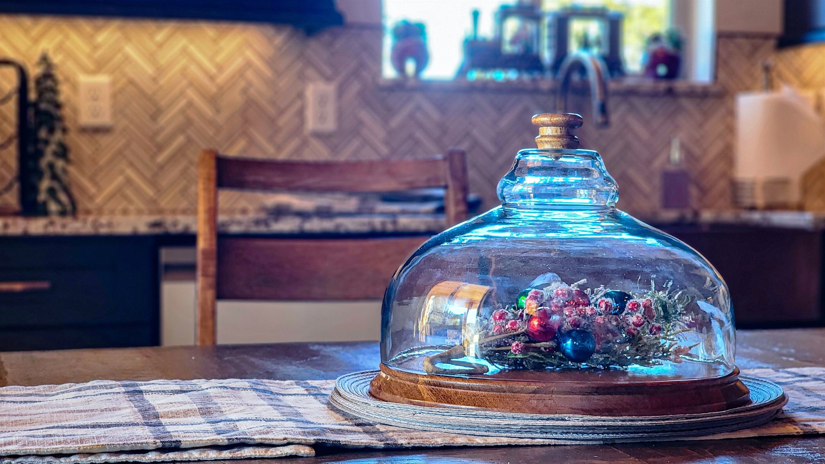 Une cloche de verre recouvre une table sur laquelle repose un set de table en fibres tressées, une chaise en bois et, en arrière-plan, un mur orné d’un motif en chevrons.