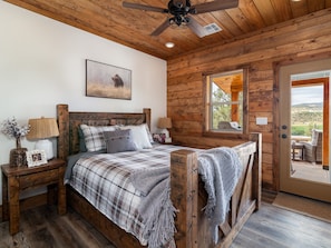 Une chambre à coucher située dans une cabane en bois, comprenant un lit, des tables de chevet, un ventilateur de plafond, une fenêtre avec vue et une porte donnant sur l’extérieur.
