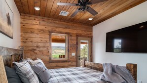 Un dormitorio de cabaña de madera con colcha a cuadros, ventilador de techo y ventana grande con vistas al exterior.