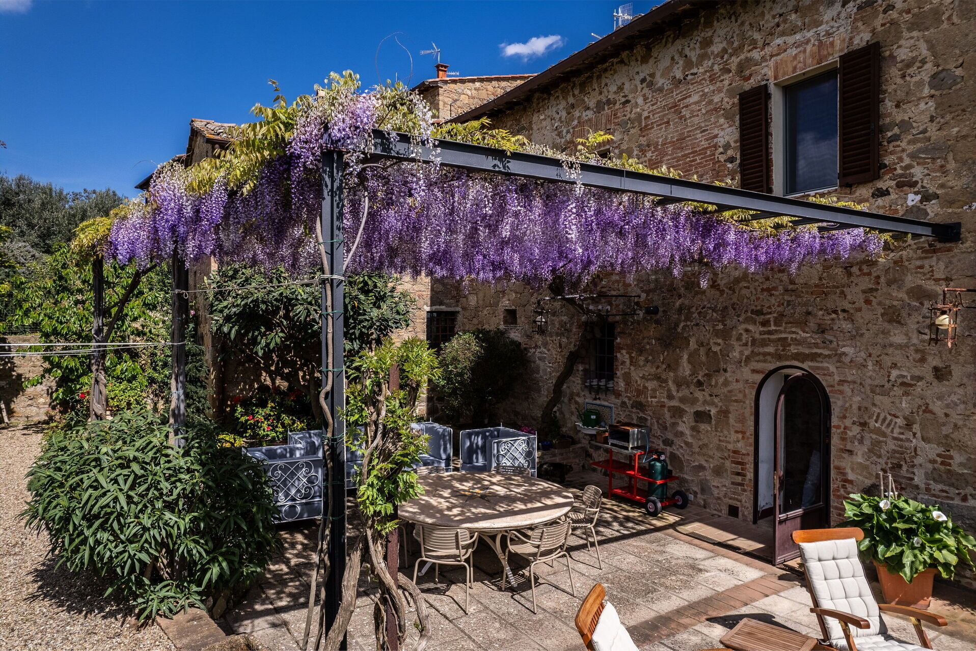 Ein rustikales Steinhaus mit einer Weinlaube, die in violette Blüten getaucht ist, mit Außenbestuhlung und einem kleinen Garten.
