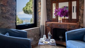 A room with a stone wall, a large window with a view of the sea and trees, a wooden cabinet with a mirror, and two blue armchairs.