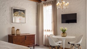 Une pièce comprenant une commode en bois, une table blanche avec des chaises, un téléviseur fixé au mur et un lustre.