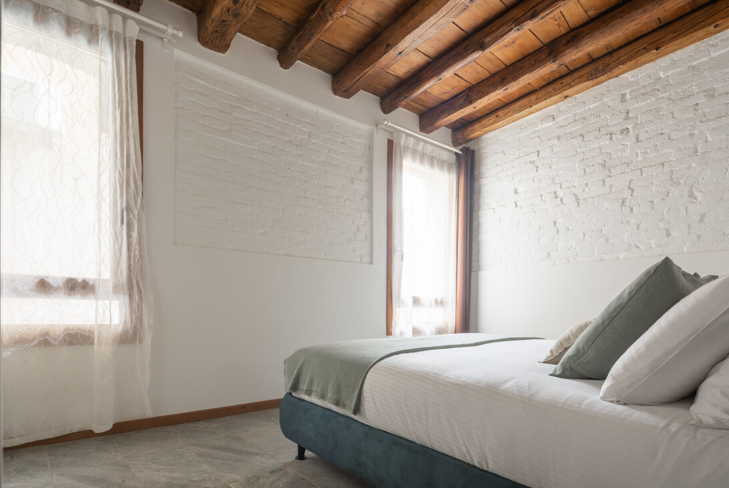 Camera da letto con un letto, travi in legno, pareti in mattoni bianchi e tende leggere.