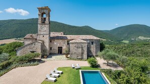 Edificio de piedra con torre de campanas, piscina y zona verde con sillones de descanso.