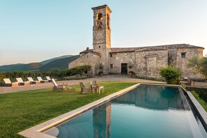 Un edificio de piedra con una torre, piscina y sillones de descanso.