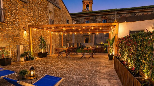 Un edificio de piedra con un patio cubierto que cuenta con mesas, sillas, plantas en macetas y un sillón azul.