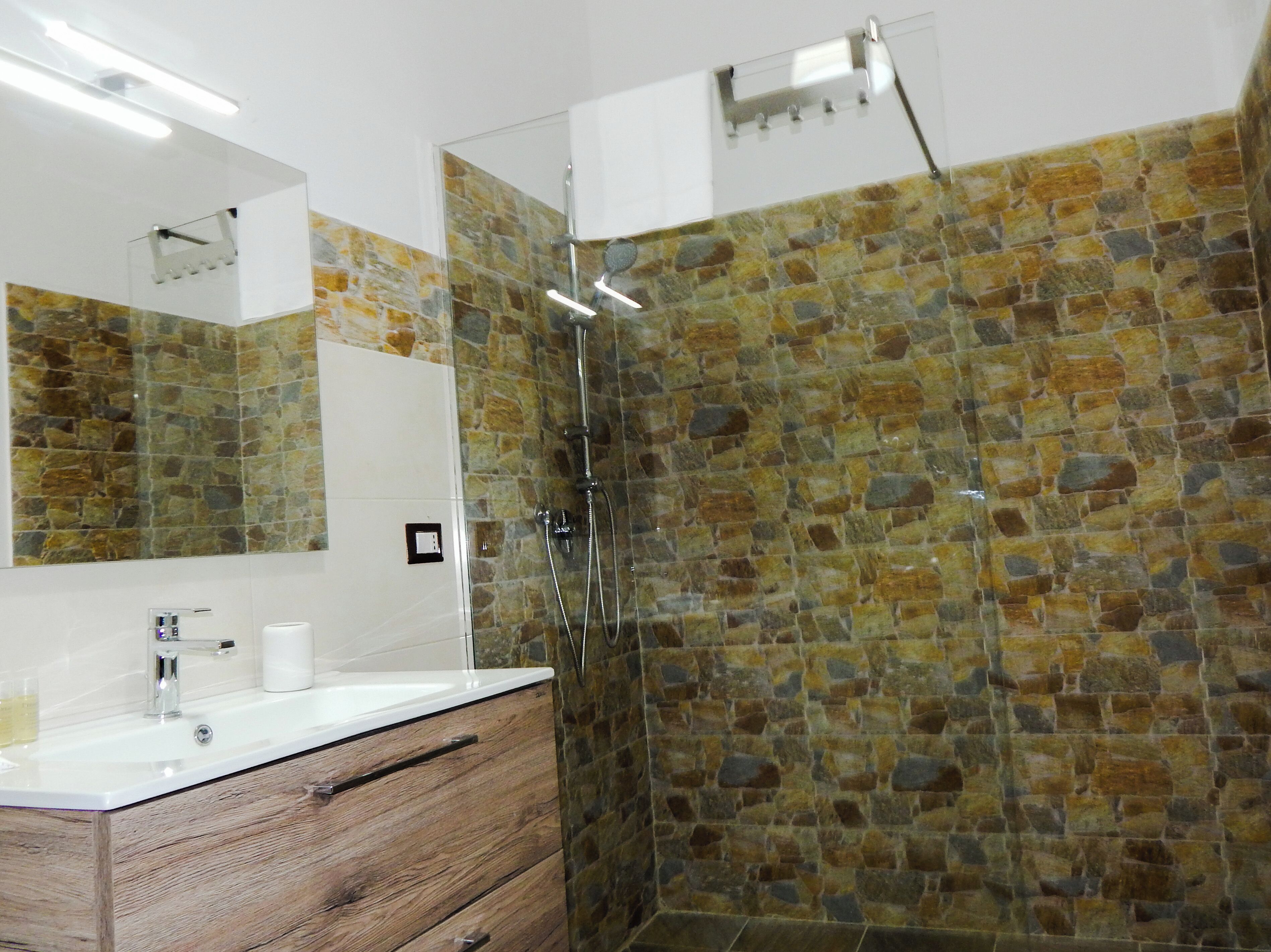 A bathroom with a stone wall shower, a wooden vanity with a mirror, and a white sink.