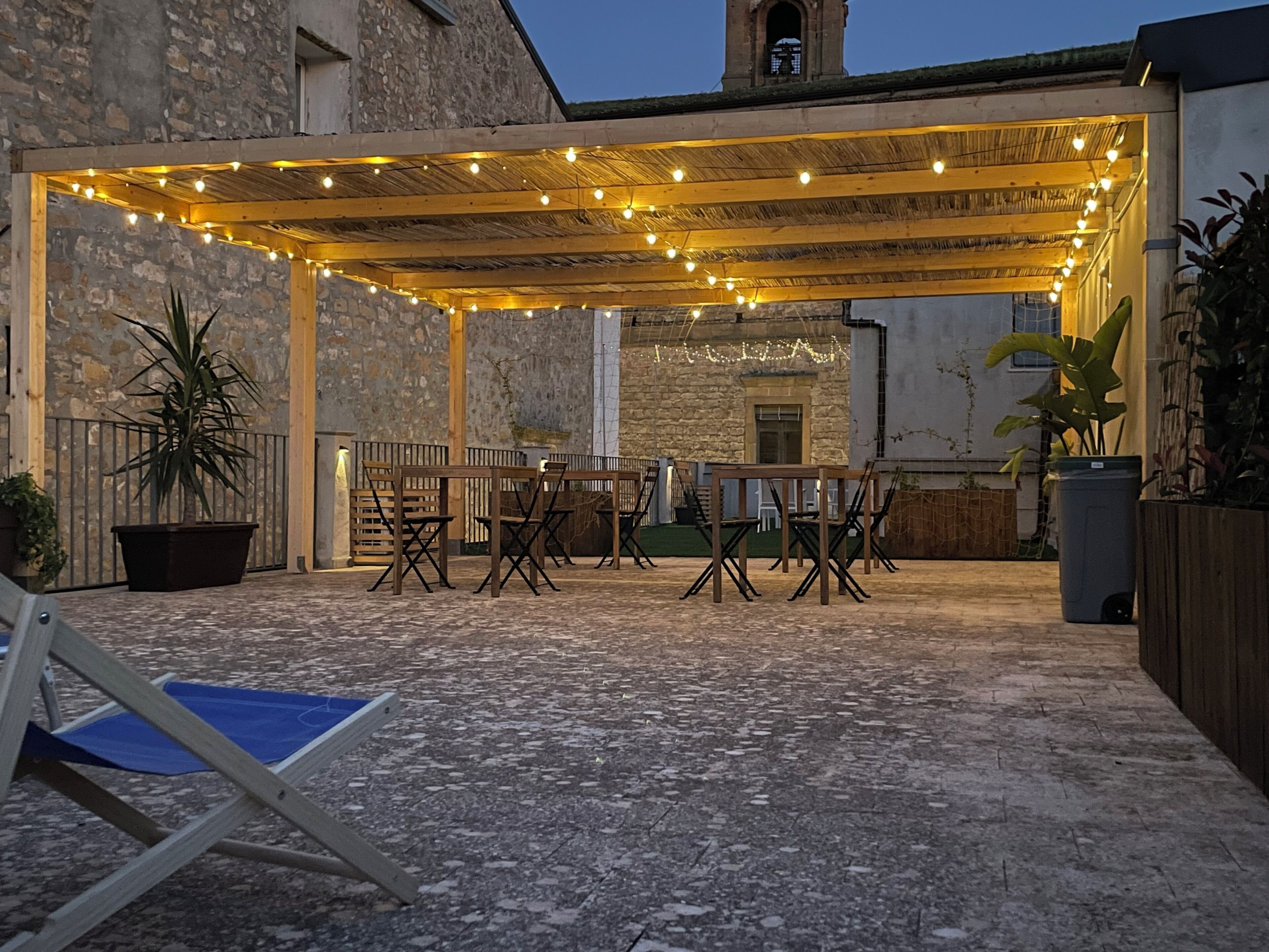 An outdoor patio with wooden tables and chairs, string lights, and potted plants.