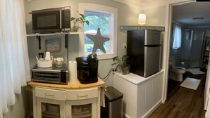Eine kompakte Küche mit Mikrowelle, Toaster, Kaffeemaschine und einem kleinen Mülleimer. Eine Seestern-Dekoration hängt am Fenster, und ein Schild über dem Kühlschrank zeigt den Text: "The Beach is My Happy Place".