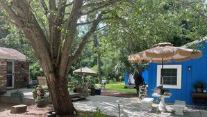 Ein Garten mit Baum, Gartenmöbeln, einem blauen Haus und einem Reetdachschirm.