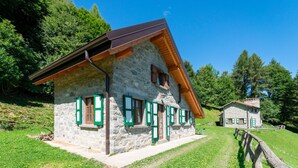 Una casa de piedra con contraventanas verdes y techo inclinado, rodeada de vegetación y una valla de madera.