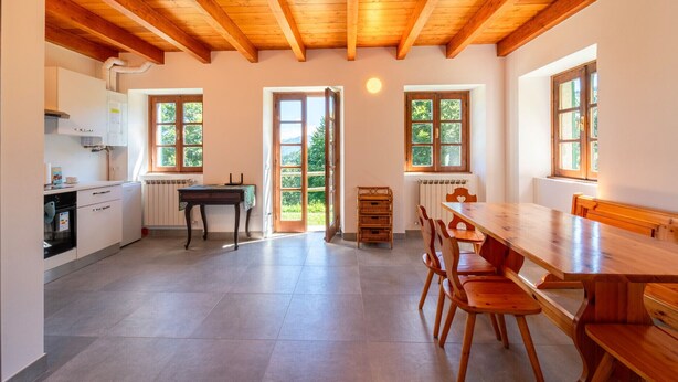 Una cocina amplia con vigas de madera, un comedor con una mesa grande y sillas, y múltiples ventanas que ofrecen vistas a la vegetación.