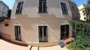 A multi-story building with balconies, a tiled ground floor, and a garden area with flowers.