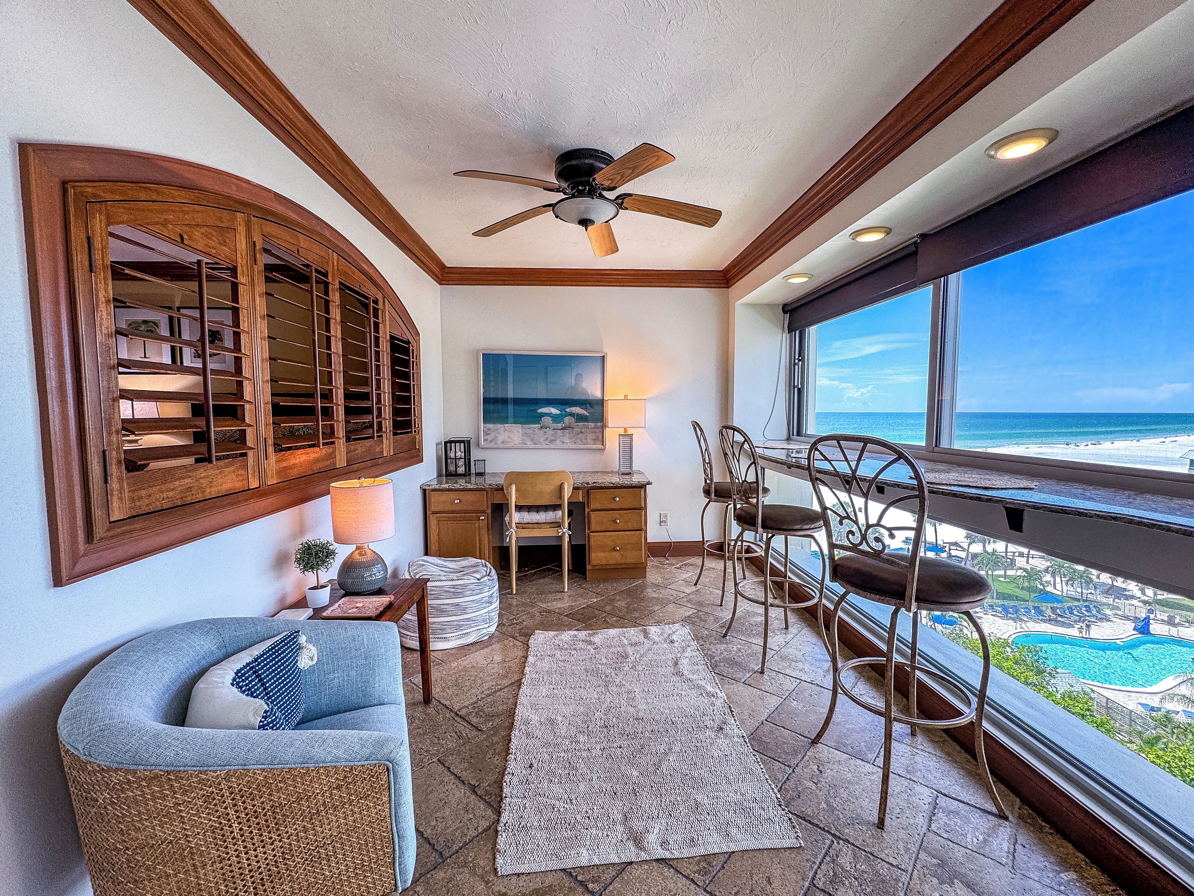 Une pièce comprenant un bureau, des chaises, un ventilateur de plafond, une grande fenêtre donnant sur la plage et un cadre de fenêtre en bois décoratif.