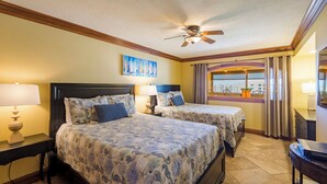 A hotel room with two beds, a ceiling fan, a painting, a window with curtains, and a desk with a lamp.