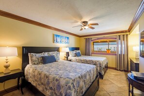 A hotel room with two beds, a ceiling fan, a painting, a window with curtains, and a desk with a lamp.