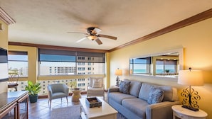 A living room with a ceiling fan, a sofa, a coffee table, a chair, a small table with a lamp, and a view of buildings through large windows. - Unit 84- 1 Bedroom 1 Bathroom Condominium with Partial North Views. (Sarasota)