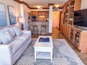 A living room with a sofa, coffee table, and a kitchen area with wooden cabinets and a TV. - Unit 84- 1 Bedroom 1 Bathroom Condominium with Partial North Views. (Sarasota)