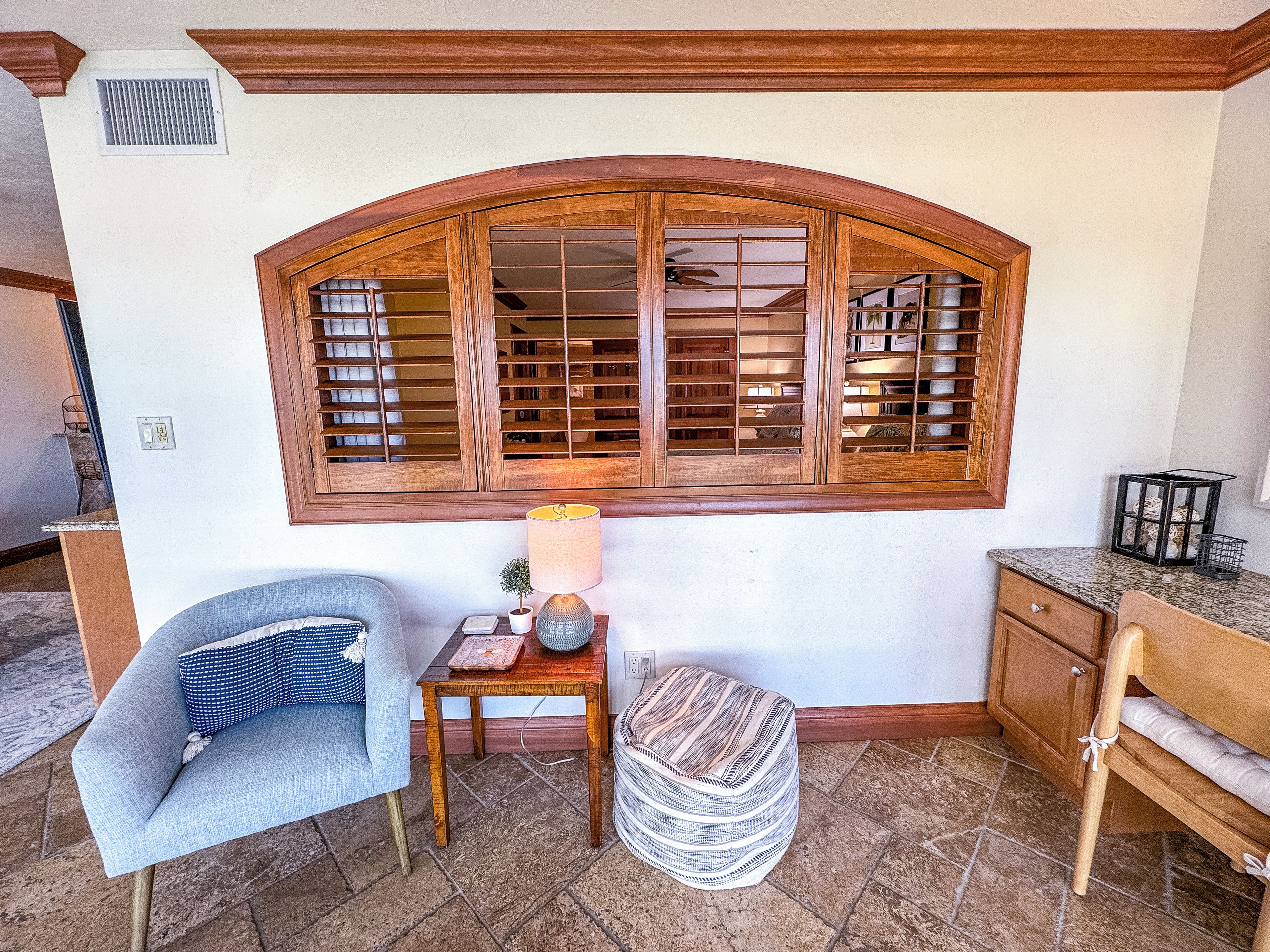 A room with a large arched window, a small table with a lamp, a chair, and a cabinet.