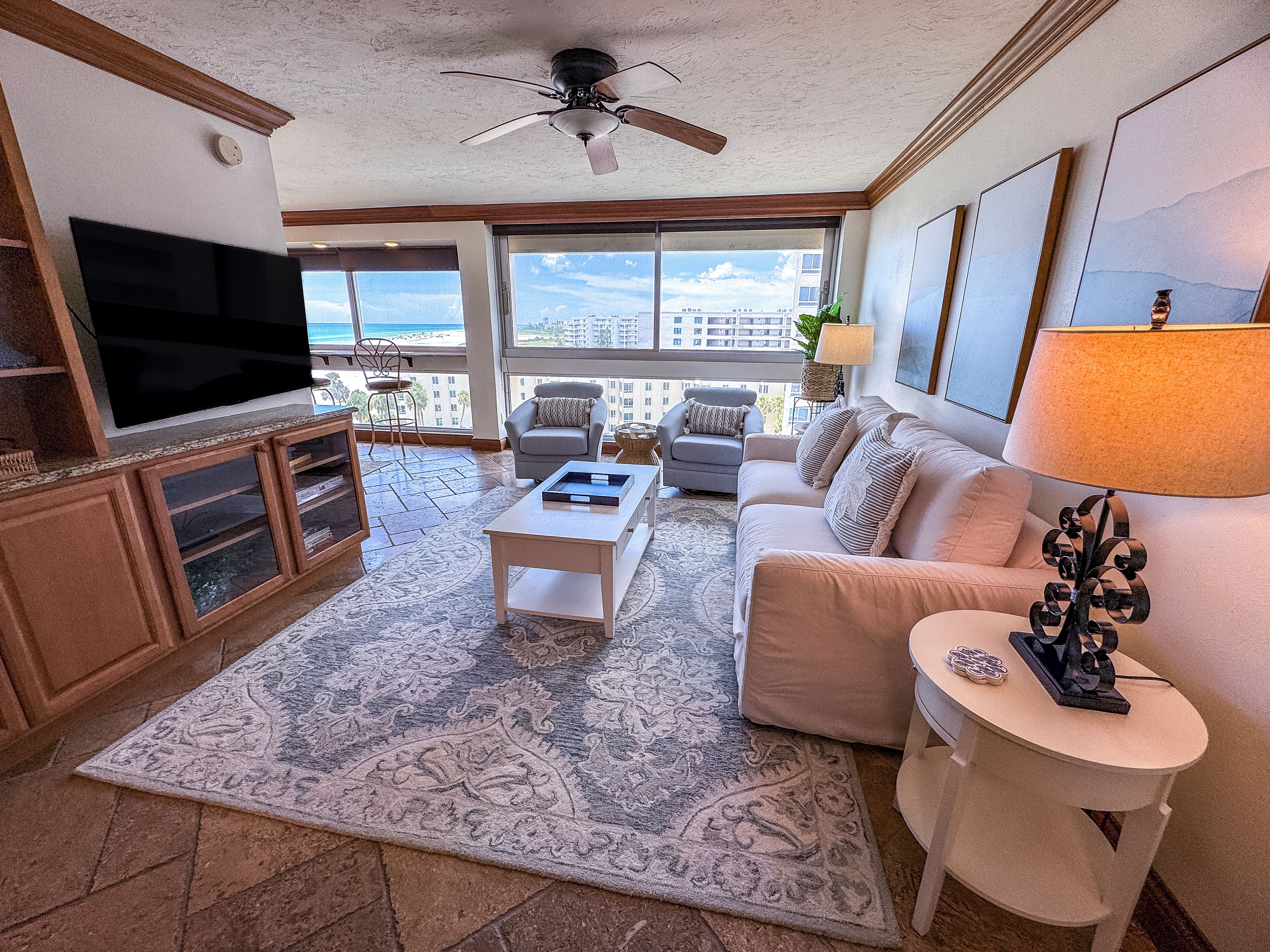 A living room with a flat-screen TV, a ceiling fan, a large window with a view, a sofa, a coffee table, and a patterned rug.