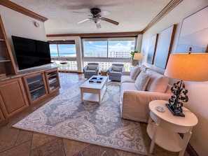 A living room with a flat-screen TV, a ceiling fan, a large window with a view, a sofa, a coffee table, and a patterned rug. - Unit 84- 1 Bedroom 1 Bathroom Condominium with Partial North Views. (Sarasota)