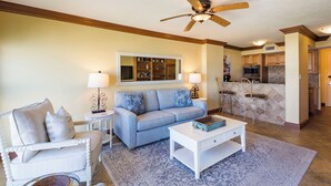 A living room with a sofa, coffee table, and chairs, a kitchen area with a bar, and a ceiling fan.