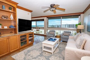 A living room with a flat-screen TV, a ceiling fan, a sofa, a coffee table, and a view of the ocean through large windows. - Unit 84- 1 Bedroom 1 Bathroom Condominium with Partial North Views. (Sarasota)