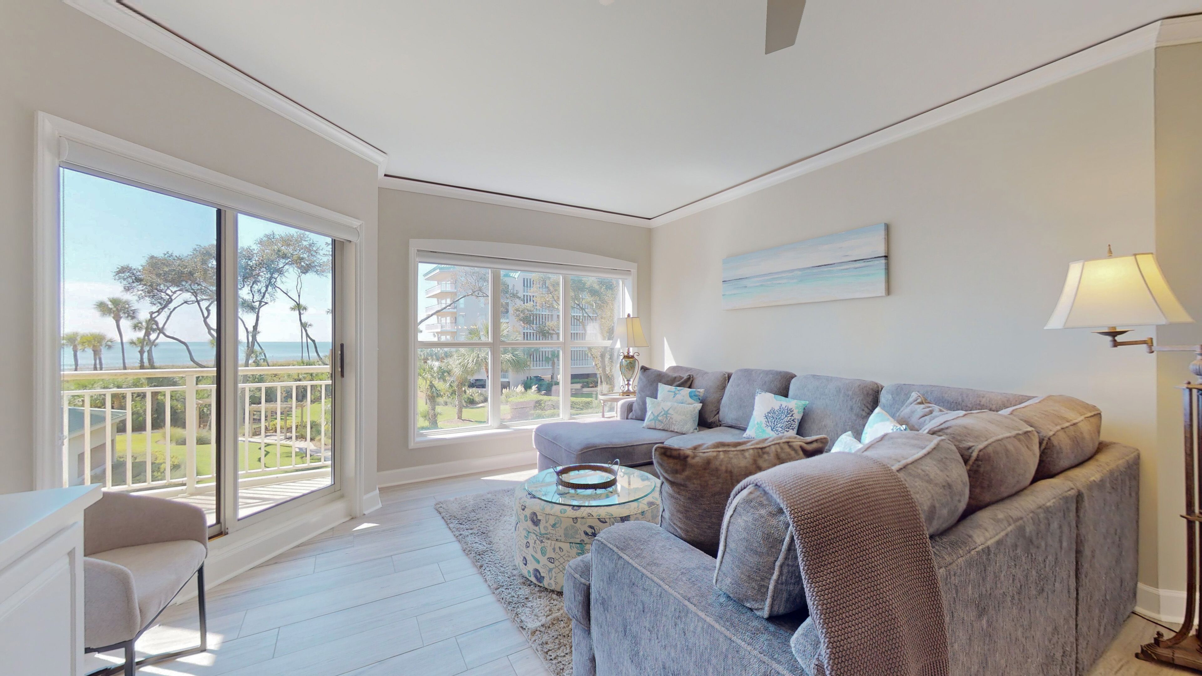 A spacious living room with a large window offering a view of a balcony and the ocean. There is a grey sofa with multiple cushions and a small round table with a decorative item.