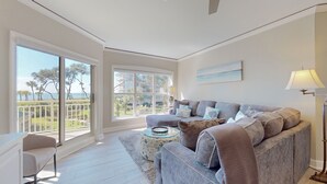 Un ampio soggiorno con una grande finestra che offre vista su un balcone e sull'oceano. C'è un divano grigio con numerosi cuscini e un tavolino rotondo con un oggetto decorativo.