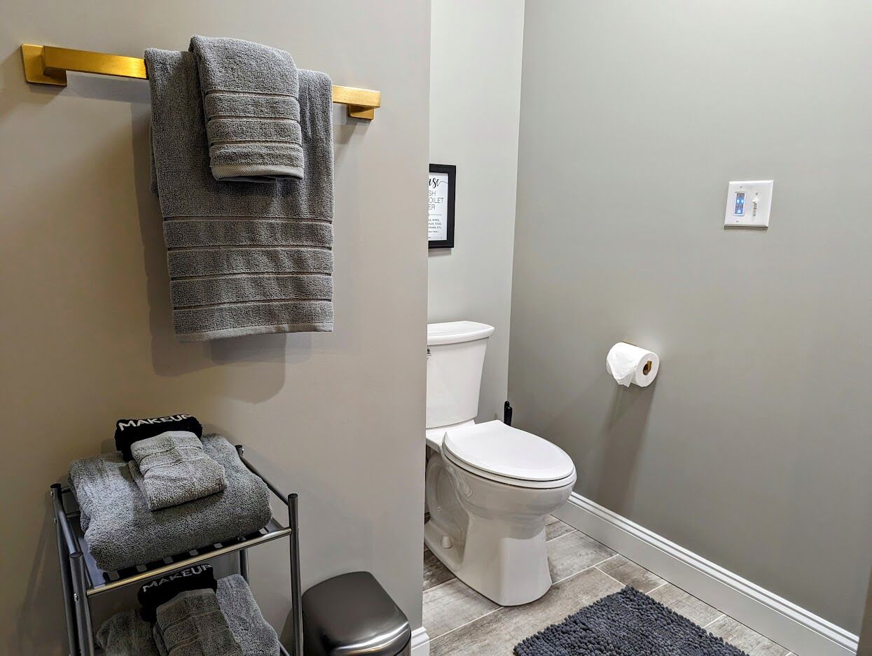 A bathroom with a toilet, a towel rack with folded towels, a trash bin, and a framed sign on the wall.