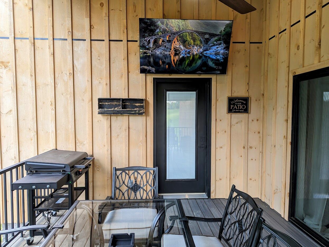 A wooden deck with a grill, two chairs, a table, and a TV mounted above a door.