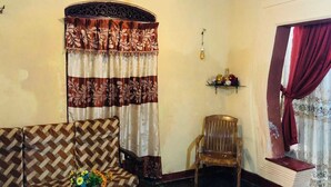 A living room with a patterned sofa, a wooden chair, a small table with a vase, and a window with curtains.