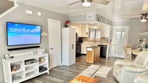 A modern living room with a flat-screen TV, a wooden coffee table, a sofa, and a kitchen area with white cabinets and a dining table. - Pet Friendly • Walk to Beach • 11 Pools • Sleeps 6 (Panama City Beach)