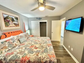 Une chambre à coucher comprenant un lit, un ventilateur de plafond, un téléviseur et une porte donnant sur une autre pièce.