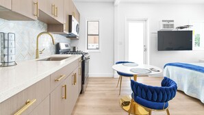 Cocina moderna con encimera blanca, detalles dorados y una estufa. Se observa un comedor con una mesa redonda y sillas azules. Al fondo, una cama con colcha azul y blanca.