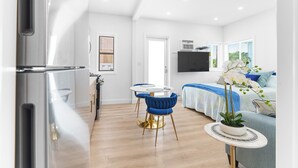 Una cocina moderna con electrodomésticos de acero inoxidable, un comedor con una mesa redonda y sillas azules, un dormitorio con una cama y un televisor, y una sala de estar con un sofá y una planta en maceta.