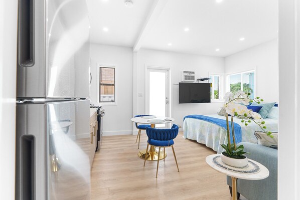 A modern kitchen with stainless steel appliances, a dining area with a round table and blue chairs, a bedroom with a bed and a TV, and a living area with a sofa and a potted plant.