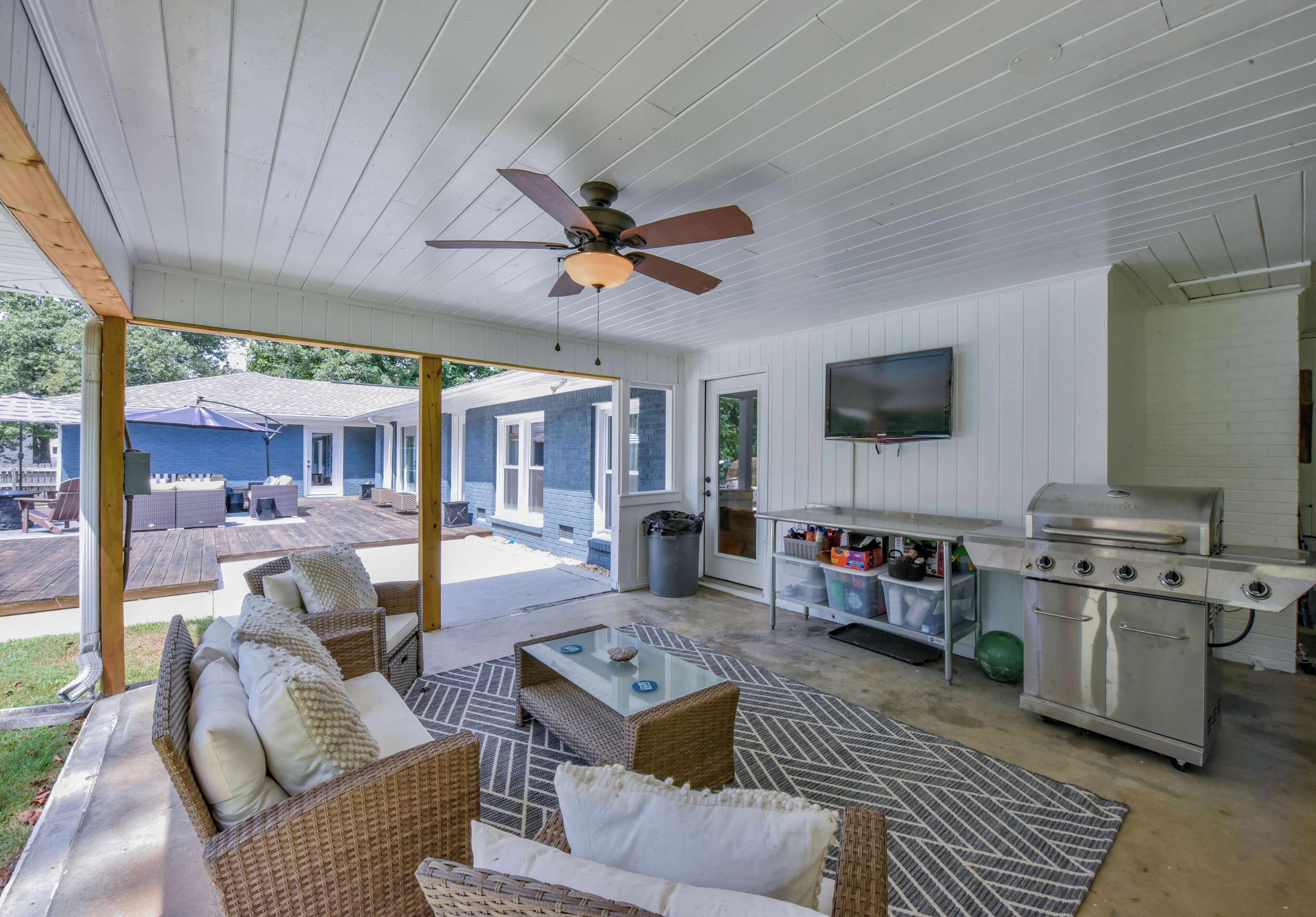 A covered patio area with wicker furniture, a glass-top table, a ceiling fan, a grill, and a television.