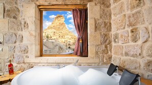 Ein Badezimmer mit Steinsäulen, einer großen Badewanne, einem Fenster mit Bergblick und einer Flasche Alkohol auf einem Holztisch.