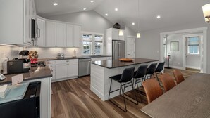 A modern kitchen with white cabinets, a large island with bar stools, a stainless steel refrigerator, and a dining area with a table and chairs. - Seabrook Play Among Stars | Hot Tub, Ocean View | Pet Friendly, 4BR, Sleeps 8 (Pacific Beach)