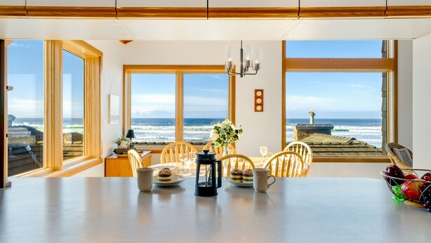 A dining area with a view of the ocean, wooden chairs, a table with a coffee pot, and a vase with flowers.