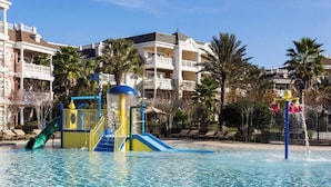 Piscina con toboganes y fuentes, rodeada de palmeras y un edificio con balcones.