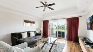 Sala de estar con un sofá, una mesa de centro de vidrio, un ventilador de techo, un televisor y un cerramiento de vidrio corredizo que da a un balcón.